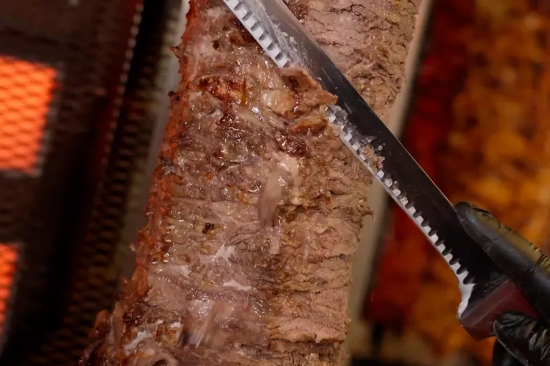 A gloved hand carving beef shawarma off the spit in front of the broiler